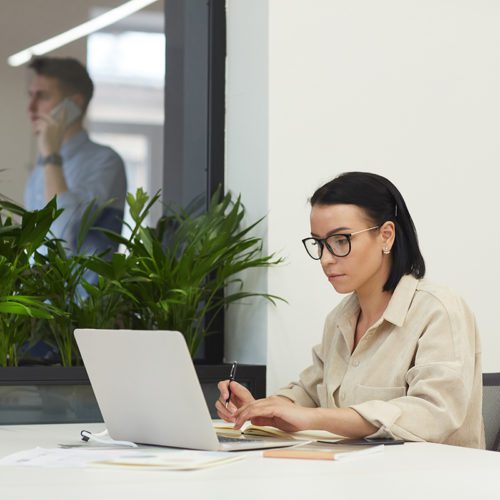 Serious manager in eyeglasses concentrating on her online work she working on laptop computer at office