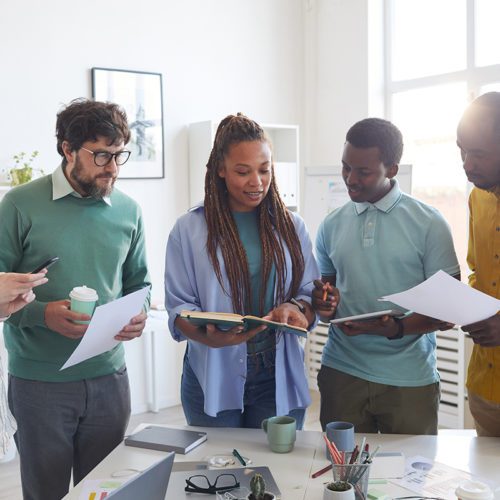 Portrait of contemporary multi-ethnic business team standing around table in office and listening to female African-American leader giving instructions, copy space