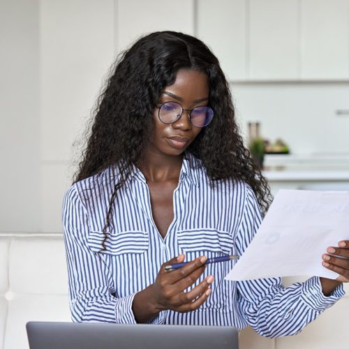 Serious young black African ethnic woman entrepreneur holding paper document checking bill, calculating taxes reading letter doing paperwork using laptop computer remote working online at home office.